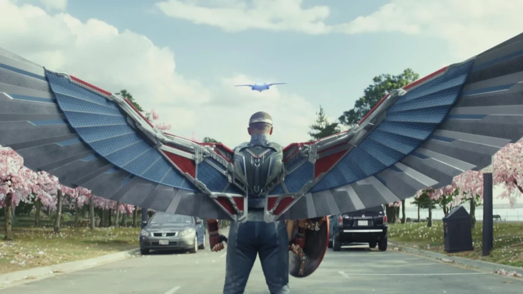 Rear view of Sam Wilson in his metallic Captain America suit with massive blue and red wings outstretched on a paved street.