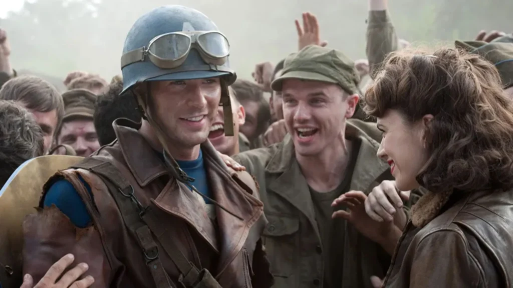 Steve Rogers smiling in his World War II Captain America field jacket and helmet among cheering soldiers and Peggy Carter.