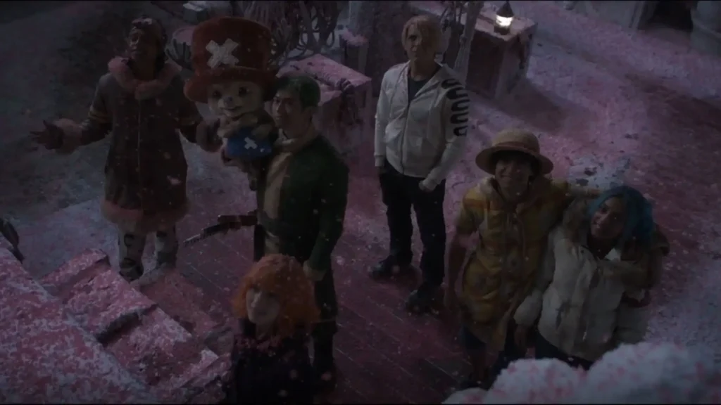The Straw Hat Pirates and their allies stand together in a snowy courtyard beneath drifting pink cherry blossom snow, looking upward with hopeful expressions outside Drum Castle. Chopper stands at the center beside Luffy, while Zoro, Sanji, Nami, Usopp, and Vivi gather around during the emotional finale of One Piece live-action Season 2, Episode 8, "Deer and Loathing in Drum Kingdom."