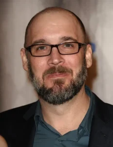 Headshot of comic book writer Ed Brubaker wearing glasses and a dark blazer smiling at an event.