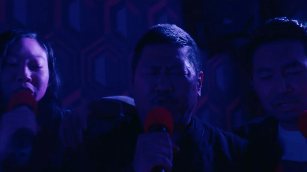 Katy, Wong, and Shang-Chi singing into microphones in a dimly lit, purple-tinted karaoke room.
