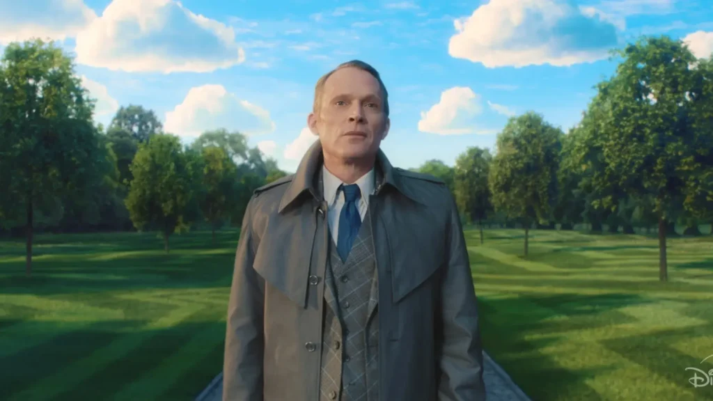 Paul Bettany as Vision in human disguise, wearing a trench coat and suit while standing on a paved path in a surreal, perfectly manicured outdoor park setting.