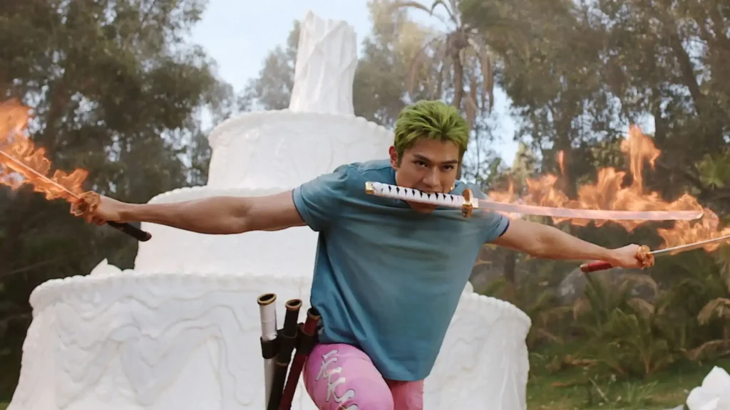 Zoro fights with his signature three-sword style, holding one katana in his mouth while wielding two flaming swords in front of a giant wax structure on Little Garden in Netflix's One Piece Season 2 Episode 5, "Wax On, Wax Off."