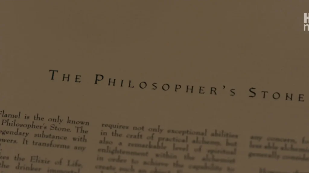 Close-up of a book page showing the heading "THE PHILOSOPHER'S STONE" and printed text detailing Nicolas Flamel.