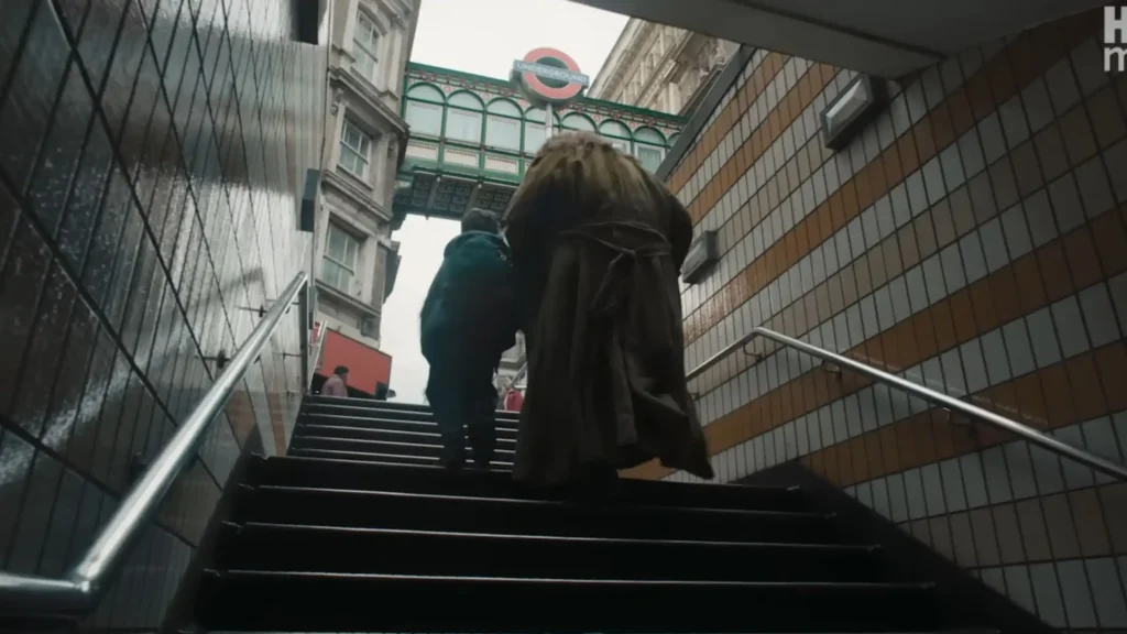 Low-angle shot of Hagrid in a long brown coat walking up metro stairs alongside young Harry toward a London Underground sign.