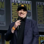 Marvel Studios President Kevin Feige wearing a black cap and blazer while speaking into a microphone on stage at San Diego Comic-Con in front of a Comic-Con logo backdrop.