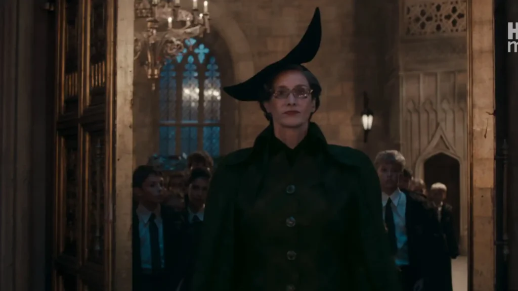 Professor McGonagall in a green cloak and pointed black hat leading a group of young first-year students into a hall.