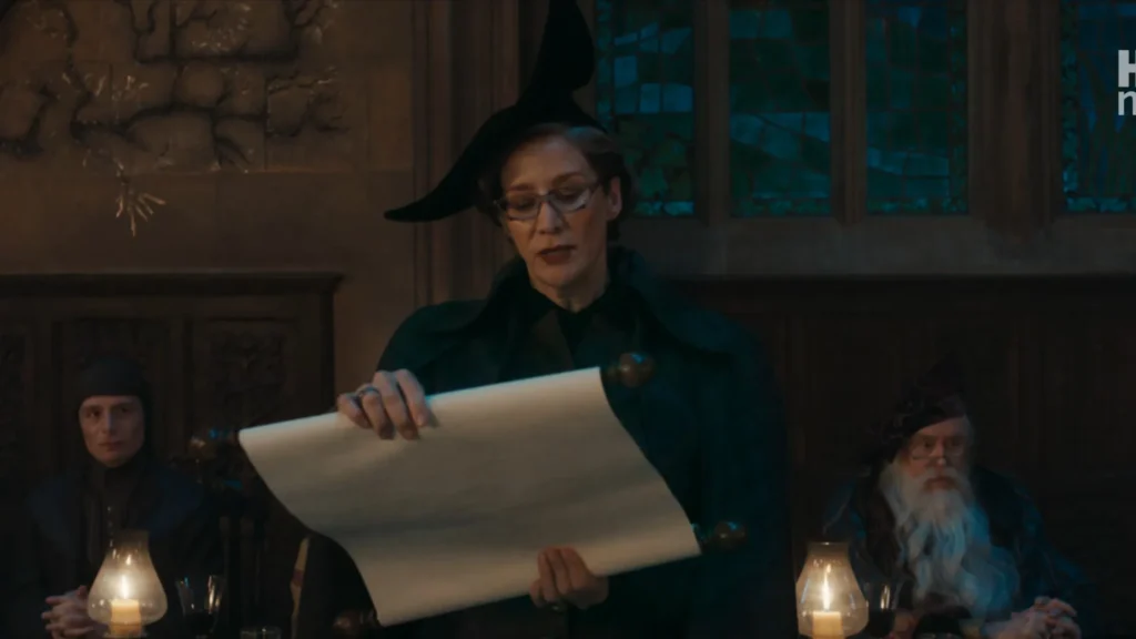 Professor McGonagall in glasses and a black witch hat unrolling a large scroll at the staff table, flanked by Quirrell and Dumbledore.