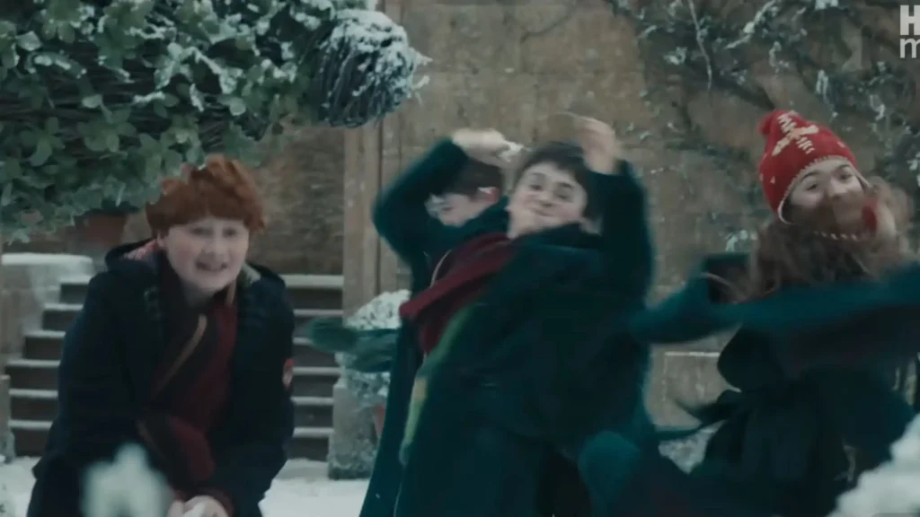 Young Ron, Harry, and Hermione wearing winter coats and scarves playing in the snow outside Hogwarts castle.