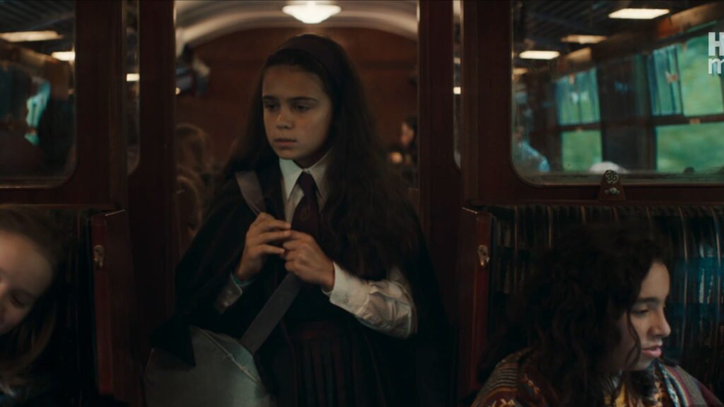 Hermione Granger in a school uniform standing in the wooden corridor of a Hogwarts Express train carriage.