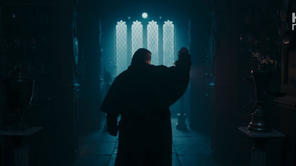 Silhouette of Argus Filch holding a raised lantern in a dark hallway lined with trophy cases and a tall arched window.
