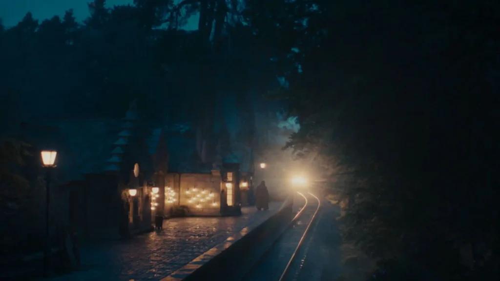 A train's bright headlight approaching a dimly lit, atmospheric stone train station platform at dusk.