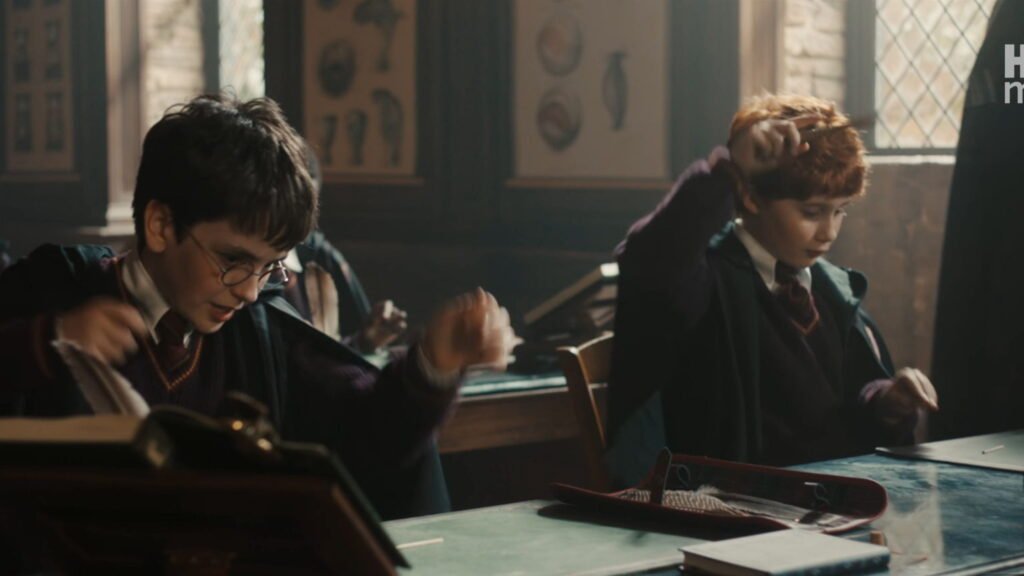 Harry Potter and Ron Weasley sitting at a classroom desk practicing waving their wands down at matchsticks.