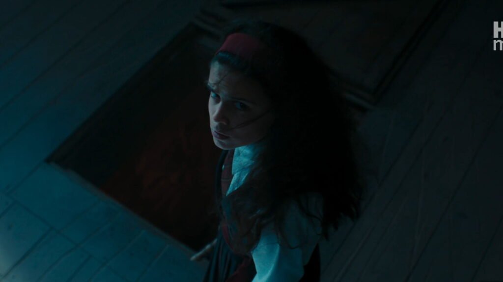 High-angle shot of young Hermione Granger wearing a red headband looking up nervously while standing next to an open wooden trapdoor.